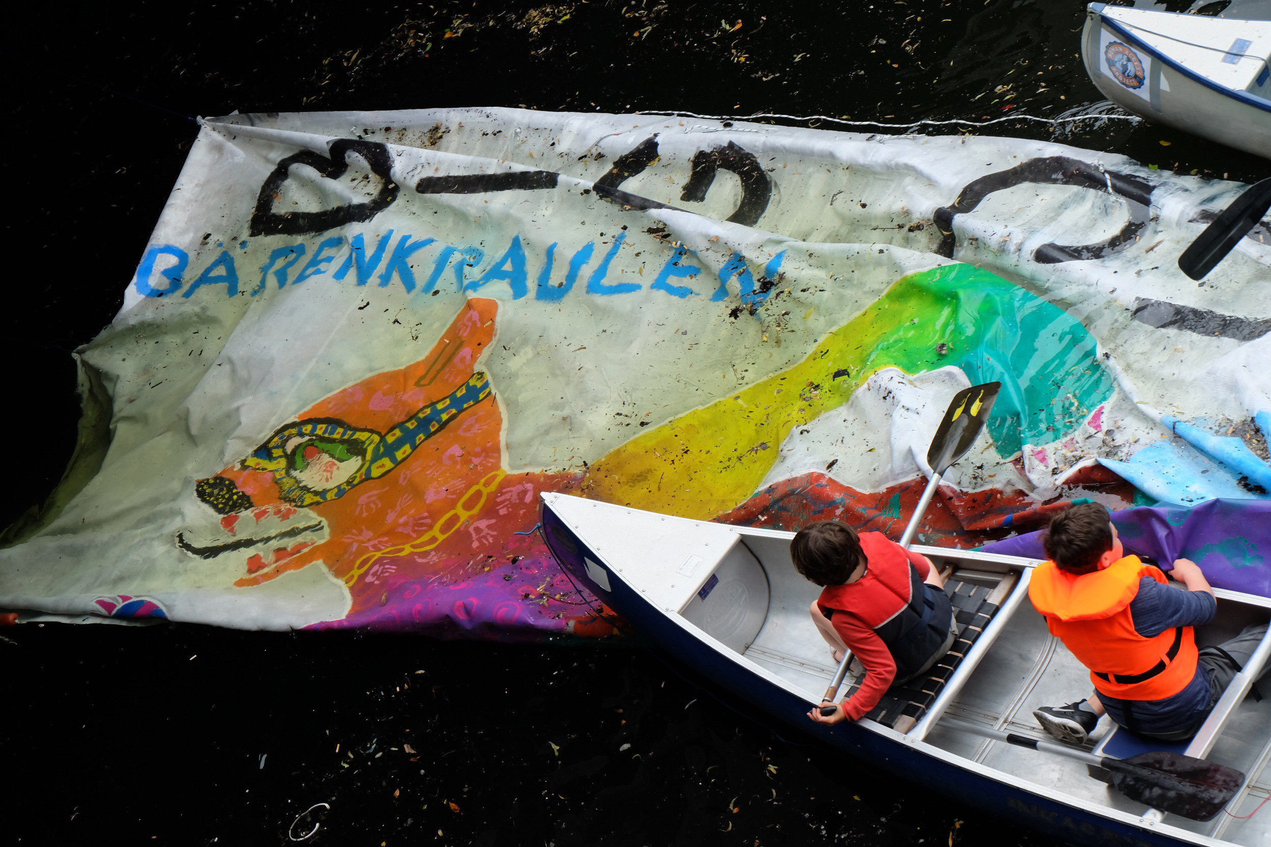 Kinder paddeln mit Kanu über ein Banner in der Spree