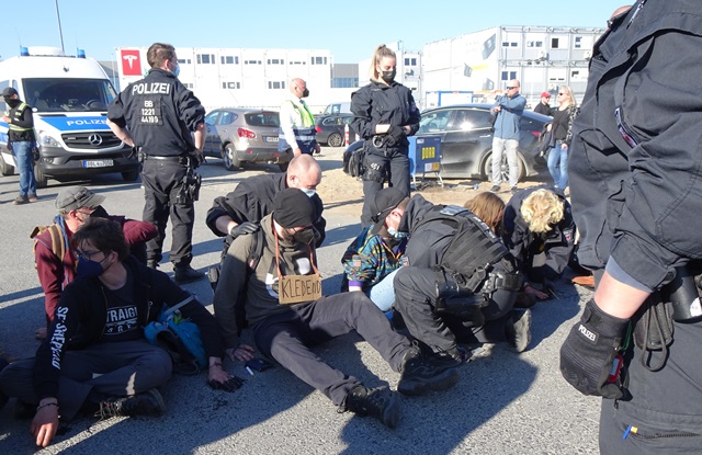 Protestaktion vor der sogenannten Gigafactory von Tesla in Grünheide bei Berlin: Einige haben sich mit den Händen am Asphalt festgeklebt, Polizei steht herum, zwei Beamte versuchen die Festgeklebten vom Boden zu lösen.